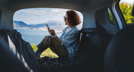 Young woman sitting in the open trunk of a car overlooking the sea with a smartphone in her hands, summer vacation and auto travel - obrazy, fototapety, plakaty