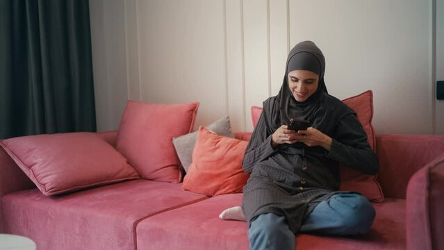Beautiful Arab female taking selfie on smartphone, sitting on couch at home