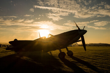 The sun rises behind a Spitfire at an airshow and shines through the cockpit glass. - obrazy, fototapety, plakaty