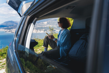 Young woman sitting in the open trunk of a car overlooking the sea with a smartphone in her hands,...