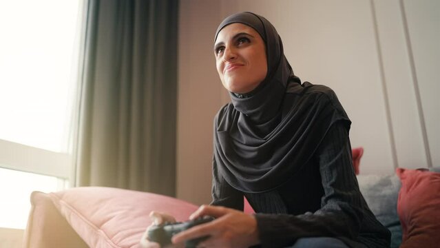Excited young woman in hijab playing video game on couch at home, bottom view