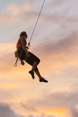 A woman is hanging from a rope, looking up at the sky. The sky is a mix of orange and pink,...
