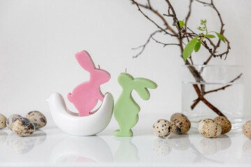 Easter concept. Colourful rabbit. Pink and light green bunnies candles. Easter quail eggs with green tree branches with leaves. Glass vase. White background, white table. Copy space.