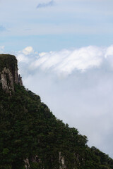 Majestic Stone Cliff in Serra do Rio do Rastro, adorned with lush green vegetation, set against a backdrop of billowing clouds.