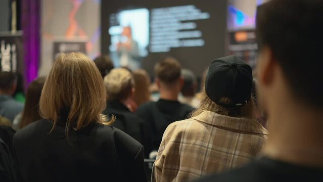 At the frontier of innovation, a presenter articulates the nuances of emerging technologies to an auditorium filled with aspiring entrepreneurs. In foreground there is blonde and girl in cap.