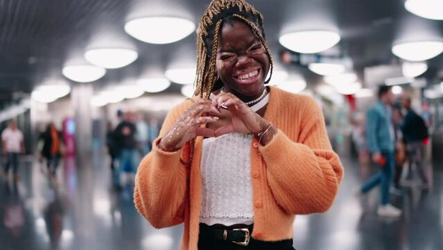 Young Black woman with vitiligo stands in busy subway station, smiling brightly and making heart shape with her hands. Female exuding positivity and warmth amidst hustle and bustle of urban life
