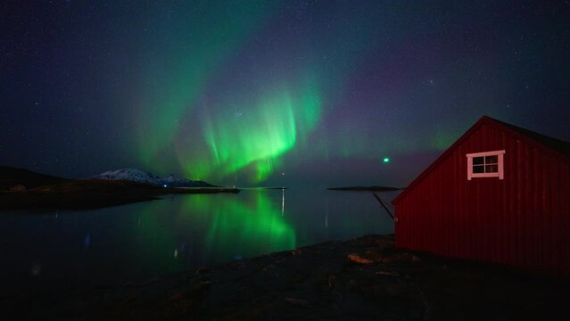 The green light from a drone can be seen hovering over the water, capturing a bright and colorful aurora reflecting in the fjords north of Mjelle, Bod&oslash;.
