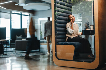 Young businesswoman speaking on smartphone , sitting inside a booth in a coworking space - 761774568