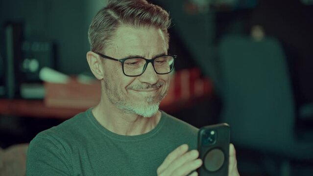 Portrait of happy confident mid adult man in glasses in office, smiling. Gray hair, bearded middle aged businessman using cell phone.