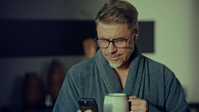 Mature man in glasses enjoying a mug of coffee while browsing internet and social media on his phone at home in morning, wearing bathrobe.
