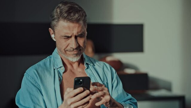 Mid adult man standing in sunlight in open shirt in the morning, checking email, news or social media on cell phone. Older male focusing, looking at smartphone.