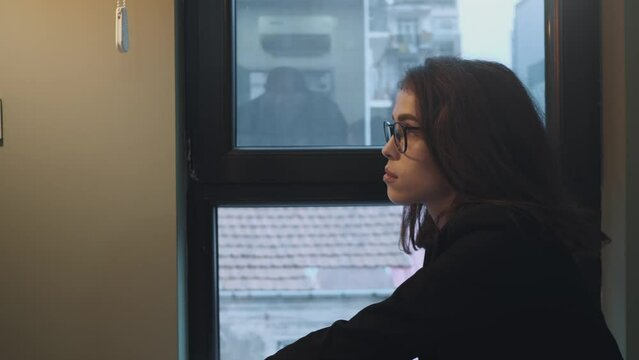 Pensive young lady with glasses looking through window, urban view.