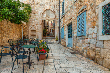 Cafe tables and chairs outside in old cozy street in the in old medieval town Hvar in outdoor restaurant with nobody, Dalmatia, Croatia. Popular travel and tourist destination on summer vacations - obrazy, fototapety, plakaty