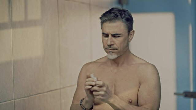Mature adult man applying cosmetics in bathroom mirror in morning. Shirtless male using anti-aging eye cream for wrinkles to stop aging.