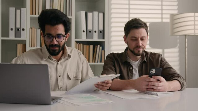 Diverse men businessmen Indian mentor salesman consult Caucasian customer explain business project on laptop in office disinterested client ignoring addicted addict with mobile phone misunderstanding