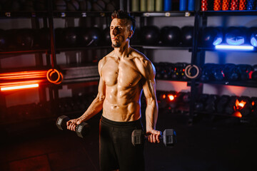 Strong shirtless bodybuilder working out biceps with dumbbells at gym.