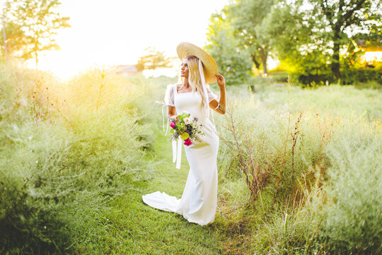 Bride in Field During Golden Hour in Early Fall in Arkansas, Sun Setting Behind Bride Wearing Wide Brim Hat Holding Wildflower Bouquet in Early Fall