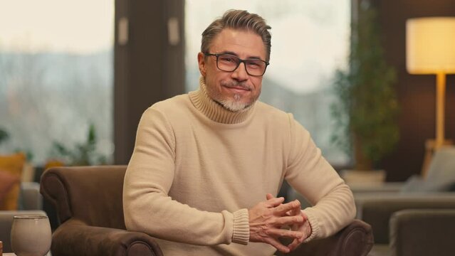 Portrait of confident middle aged man sitting at home, wearing turtleneck pullover and glasses. Mid adult older male in cosy living room, happy, smiling.