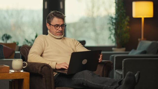 Middle aged man sitting in comfort in living room working on laptop computer in home office. Businessman managing business online. Portrait of confident older male, happy, smiling.