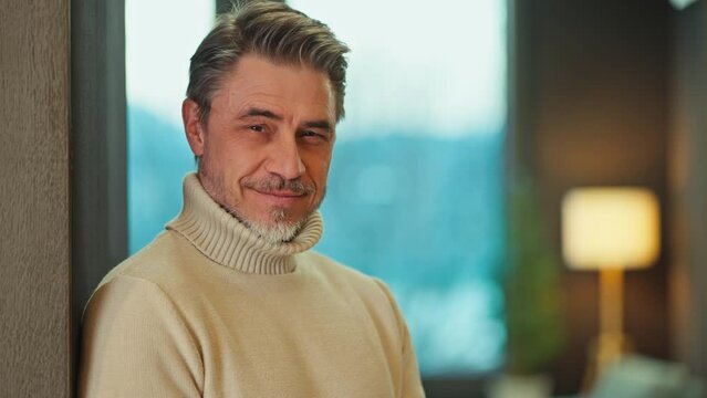 Portrait of confident middle aged man standing at home, wearing turtleneck pullover. Mid adult older male in cosy living room, happy, smiling.