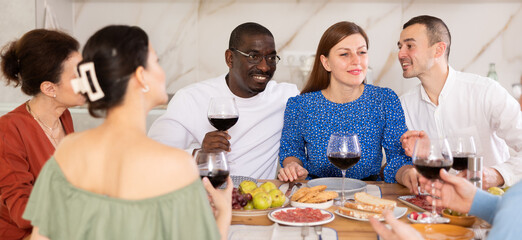 Merry male and female friends having lunch with wine and talking warmly in comfort of their house