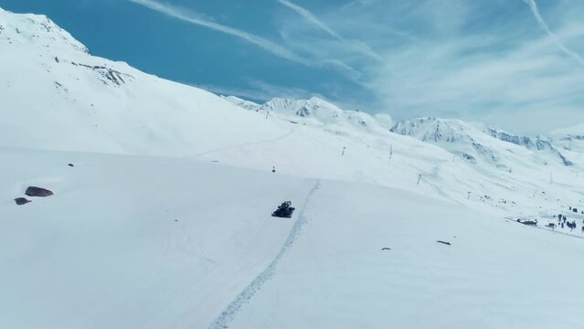 Drone follows snowcat plowing snow on ski slopes of ski resort on sunny day. Beautiful mountain landscape in Austrian Alps. Snowcat groomer prepares piste for skiing