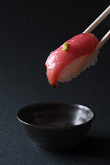 黒い背景の前でで醤油をつけ、わさびが乗ったまぐろの寿司を箸で持ち上げている