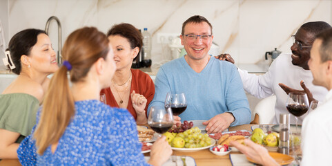 Ethnically diverse group of friends, adult women and men, sharing laughter and conversation over dinner with wine at cozy home setting..