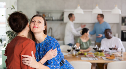 Relaxed and joyful dinner party scene at home, with smiling woman hosting friends, giving greeting kiss on cheek to bestie