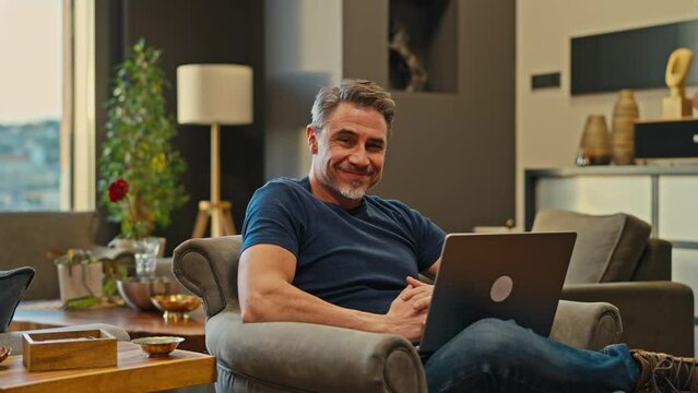 Casual mid adult man with laptop computer in home office, banking online, remote working. Portrait of happy older bearded guy smiling. Businessman, entrepreneur managing business on internet.