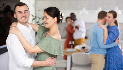 Fototapeta na wymiar Smiling young Asian woman in olive dress enjoying slow dance with husband at relaxed friendly home gathering..