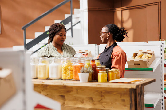Female merchant showcasing her products in zero waste marketplace venue, selling natural items to interested customer. Local vendor trading eco friendly bio food items during event to client.