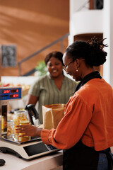 Female customer at a grocery store checkout counter with a smiling storekeeper weighing products on digital scale. Friendly vendor measuring weight of bio food items while client waits at desk.