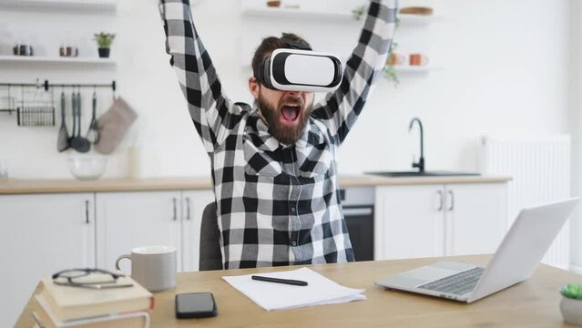 Joyful bearded man wearing VR headset and gesturing from happiness while working distance at kitchen. Caucasian technology manager satisfied with new modern gadget.