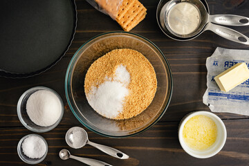 Graham Cracker Crust Ingredients with a Tart Pan: Graham cracker crumbs and sugar in a mixing bowl surrounded with other ingredients and kitchen tools