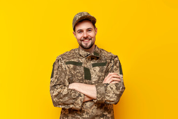 young Ukrainian army soldier in pixel camouflage uniform stands with his arms crossed on a yellow...
