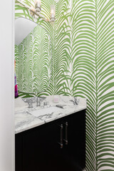 A bathroom with green and white wallpaper, a black cabinet, marble countertop, and light mounted above the mirror.