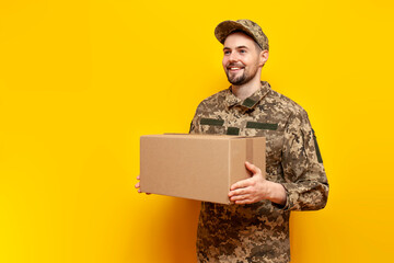 Ukrainian army soldier in pixel camouflage uniform holds a box on a yellow isolated background,...