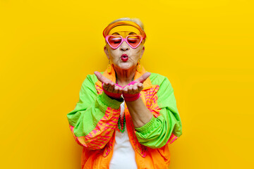 funny crazy granny in hipster clothes glasses and cap sends an air kiss on a yellow isolated...