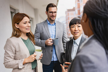 A cheerful, multiethnic group of business people stand on an office balcony, taking a coffee break and engaging in lively dialogue