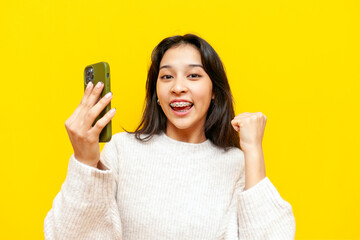 young asian woman with braces using smartphone and winning on yellow isolated background, korean...