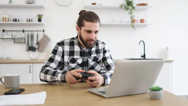Portrait of young Caucasian man using wireless joystick and laptop sitting at wooden table at kitchen. Male freelancer taking break and plays video games while working during evening time.
