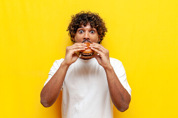 young shocked indian man eating delicious burger and surprised on yellow isolated background, curly...