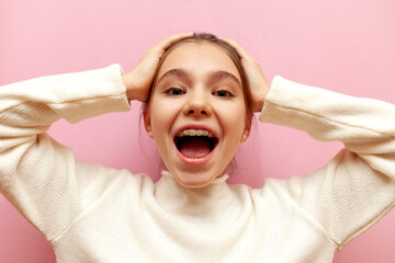 shocked teenage girl with braces screams and holds her head over pink isolated background,...