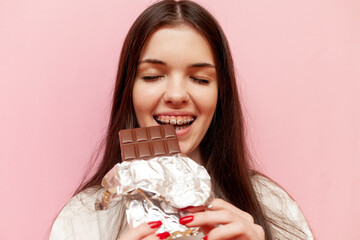 cheerful woman with braces eats chocolate and smiles on pink isolated background, young girl bites sweet food, close-up