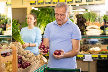 Busy focused casual elderly man buying local red onions for salad during shopping in grocery store - 761864314