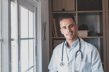 Headshot portrait of smiling young male GP or physician in white medical uniform and glasses look...