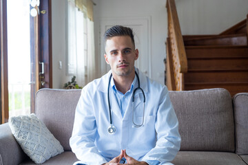 Portrait of smiling caucasian young male doctor at home in white medical uniform and stethoscope sit on the sofa look at camera, happy positive man GP or physician posing at workplace. - Powered by Adobe