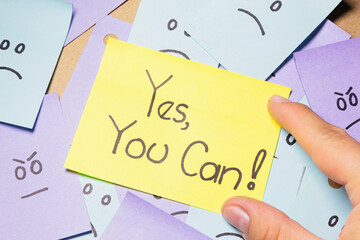 A person holds a sticky note with the message ¨Yes, you can!¨ over a pile of sticky notes with sad and angry faces. Motivational message. - obrazy, fototapety, plakaty