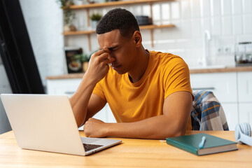 Depressed sad Latin man working at home, using laptop, eyes closed, having migraine, having bad news - obrazy, fototapety, plakaty